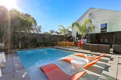 Sunlit backyard pool oasis with a rectangular pool, shallow tanning ledge featuring orange in-water loungers and round white side tables with drinks, palm trees, yellow outdoor dining chairs and wicker lounge seating beside a white house.