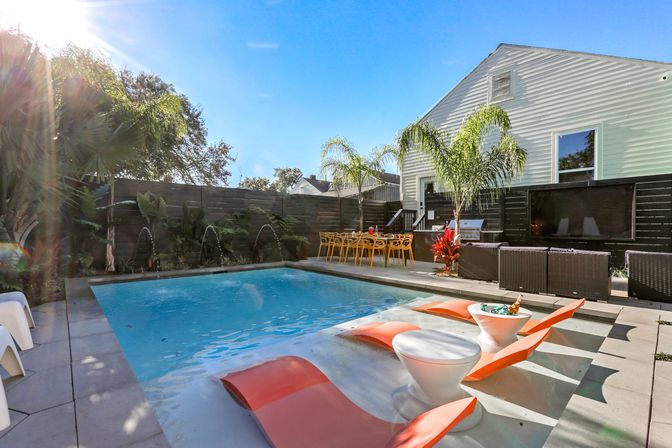 Sunlit backyard pool oasis with a rectangular pool, shallow tanning ledge featuring orange in-water loungers and round white side tables with drinks, palm trees, yellow outdoor dining chairs and wicker lounge seating beside a white house.