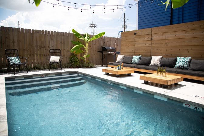 Sunny backyard pool oasis with blue water, concrete deck, long wooden sofa with patterned pillows, low coffee tables, string lights, banana plant and a grill by a privacy fence.