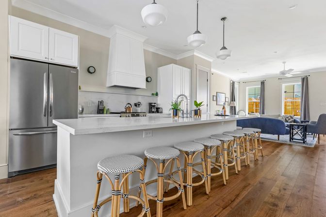 Bright open-concept kitchen and living area with a long white marble island, row of woven bistro bar stools, stainless steel French-door fridge, pendant lights, hardwood floors, and a cozy blue sofa in the adjacent seating area.