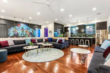 Spacious modern communal lounge with open kitchen — large dark sectional sofas with colorful pillows, round coffee tables on a pale circular rug, glossy hardwood floors, pendant lights above a bar island with metal stools, stainless refrigerator and a vibrant wall mural.