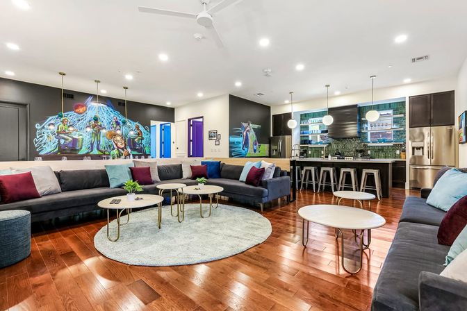 Spacious modern communal lounge with open kitchen — large dark sectional sofas with colorful pillows, round coffee tables on a pale circular rug, glossy hardwood floors, pendant lights above a bar island with metal stools, stainless refrigerator and a vibrant wall mural.