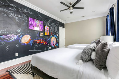 Modern urban bedroom with two white beds and decorative pillows, a bold black feature wall featuring a vibrant music-themed mural of drums, feathers and a city skyline, framed art, ceiling fan, navy curtains and a striped rug on hardwood floors.