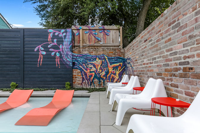 Vibrant outdoor pool patio with shallow pool and orange in-water loungers, white modern lounge chairs and red side tables against a brick wall painted with a colorful tree-and-roots mural.