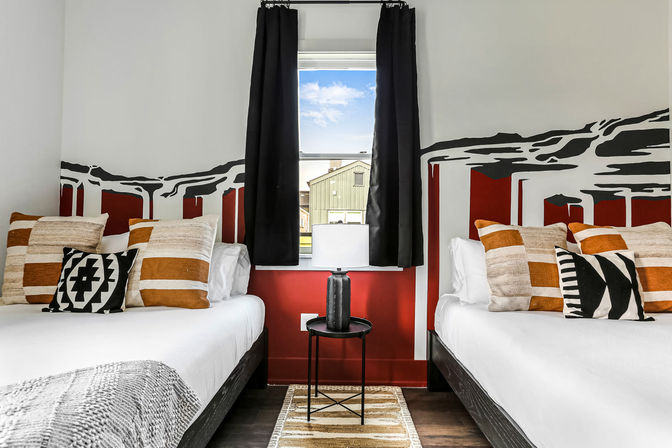 Cozy modern twin bedroom with two beds, bold red-and-white mural accent wall, black curtains, patterned throw pillows, round side table with lamp and window showing blue sky.