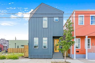 Modern narrow blue-gray townhouse with a yellow front door beside a coral-colored house on an urban street, small tree and clear blue sky