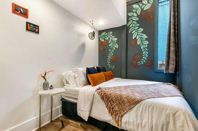 Cozy modern small bedroom with white bedding, orange throw and pillows, navy accent wall featuring a green leafy mural, small marble side table with vase, and a window with patterned curtain.