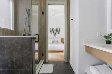 Modern bathroom with glass walk-in shower, dark tile floor, floating wood vanity with white countertop and plant, looking through doorway into a sunlit minimalist bedroom with white bedding and macramé wall hanging