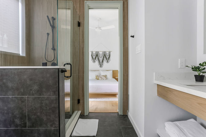 Modern bathroom with glass walk-in shower, dark tile floor, floating wood vanity with white countertop and plant, looking through doorway into a sunlit minimalist bedroom with white bedding and macramé wall hanging