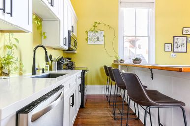 Sunny yellow kitchen with white cabinets and quartz countertop, stainless steel sink and dishwasher, black pull-down faucet, microwave and stove; wooden breakfast bar with three brown leather stools, hardwood floors, large window and trailing houseplants.