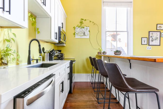 Sunny yellow kitchen with white cabinets and quartz countertop, stainless steel sink and dishwasher, black pull-down faucet, microwave and stove; wooden breakfast bar with three brown leather stools, hardwood floors, large window and trailing houseplants.