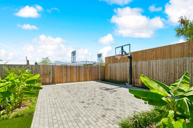 Sunlit residential backyard basketball court with gray paver patio, adjustable hoop, wooden privacy fence, tropical banana plants and a distant steel bridge under a bright blue sky.
