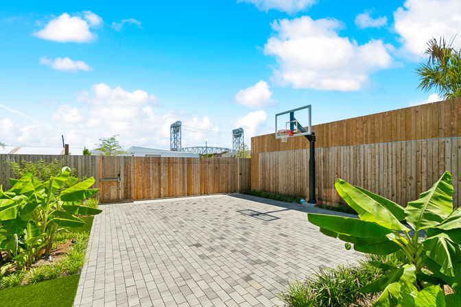 Sunlit residential backyard basketball court with gray paver patio, adjustable hoop, wooden privacy fence, tropical banana plants and a distant steel bridge under a bright blue sky.