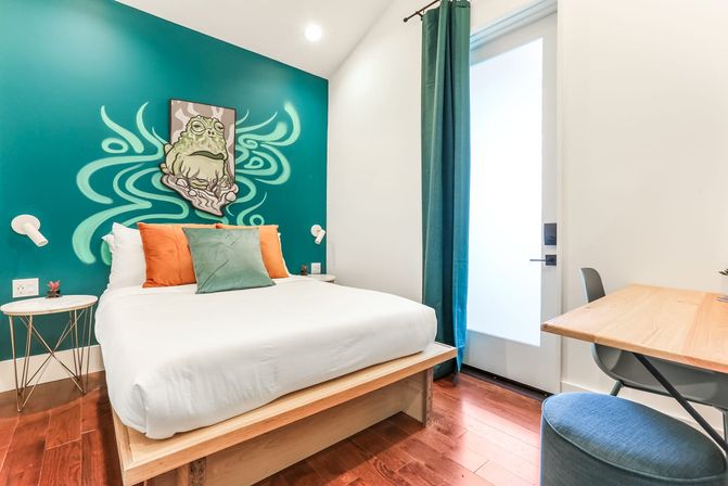 Modern bedroom with teal accent wall featuring a playful frog mural, white-lined queen bed on a wooden platform with orange and green pillows, hardwood floors, desk and teal curtain letting in soft light