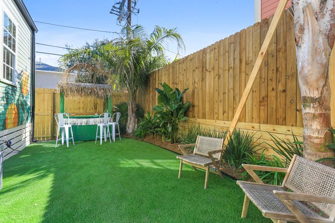 Sunny small urban backyard patio with artificial turf, wooden privacy fence, palm trees, tiki-style outdoor bar with white bar stools and woven lounge chairs.
