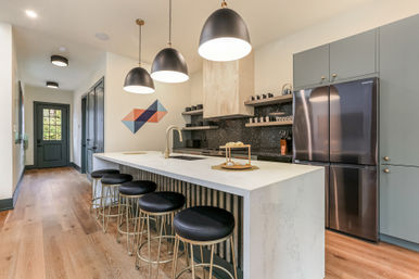 Contemporary open-concept kitchen with white marble island, five black leather bar stools with gold bases, three oversized black pendant lights, stainless French-door fridge, wood floors, geometric wall art and open shelving.