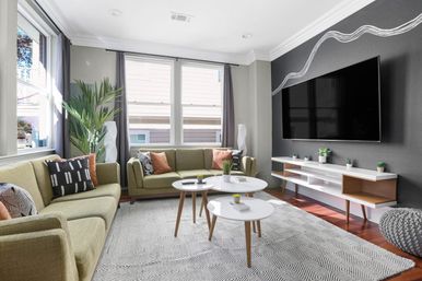 Sunlit mid-century modern living room with olive-green sofas, large windows with curtains, dark accent wall with wall-mounted TV, three round white nesting coffee tables, geometric area rug, and potted plants.