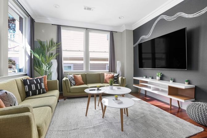 Sunlit mid-century modern living room with olive-green sofas, large windows with curtains, dark accent wall with wall-mounted TV, three round white nesting coffee tables, geometric area rug, and potted plants.