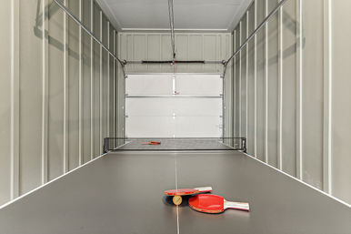 Ping-pong table set up inside a narrow garage recreation space, black table with net and two red paddles plus a ball in the foreground, white roll-up garage door at the far end.
