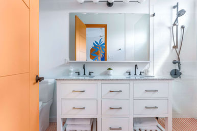 Bright modern bathroom with white double-sink vanity and speckled countertop, matte black faucets and shower fixtures, orange door, orange hex mosaic floor tiles, glass walk-in shower, and large mirror reflecting a blue-and-orange plant mural.