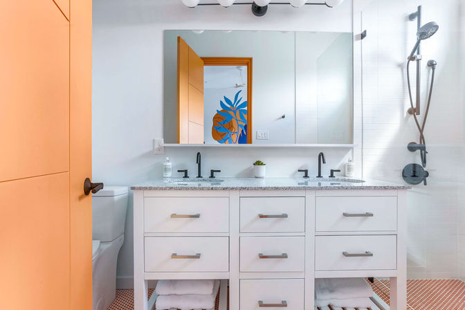 Bright modern bathroom with white double-sink vanity and speckled countertop, matte black faucets and shower fixtures, orange door, orange hex mosaic floor tiles, glass walk-in shower, and large mirror reflecting a blue-and-orange plant mural.