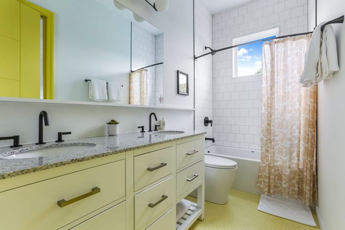 Bright modern bathroom with double-sink vanity and granite countertop, matte black faucets, subway-tiled tub-shower, patterned shower curtain and cheerful yellow floor accents