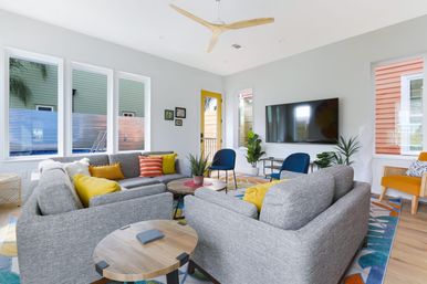 Bright, sunny modern living room with gray sectional sofas around round wooden coffee tables, yellow and orange throw pillows, blue accent chairs, wall-mounted TV, ceiling fan, indoor plants and a colorful patterned rug