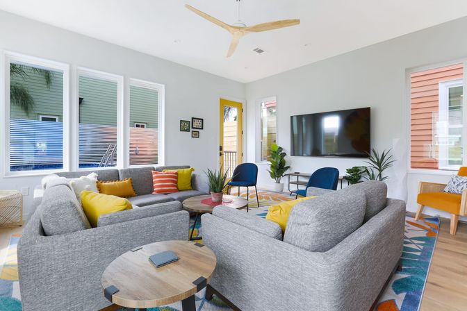 Bright, sunny modern living room with gray sectional sofas around round wooden coffee tables, yellow and orange throw pillows, blue accent chairs, wall-mounted TV, ceiling fan, indoor plants and a colorful patterned rug