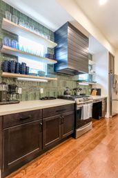 Sleek modern kitchen with glossy green tile backsplash, floating oak shelves displaying blue glassware and dishes, dark wood lower cabinets, stainless steel range and refrigerator, and warm hardwood floors.