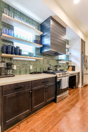 Sleek modern kitchen with glossy green tile backsplash, floating oak shelves displaying blue glassware and dishes, dark wood lower cabinets, stainless steel range and refrigerator, and warm hardwood floors.