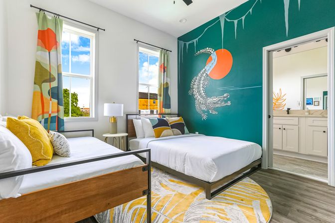 Bright modern bedroom with two beds, sunlit windows, colorful patterned curtains, playful white alligator mural and orange sun on a teal accent wall, round yellow rug and doorway to an ensuite bathroom.