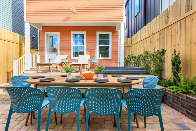 Inviting sunny backyard patio with paver flooring, long wooden dining table set with black plates and potted plants, eight turquoise chairs, coral-orange house facade, lounge chairs, hot tub, wooden privacy fence and greenery