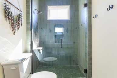 Sunlit modern bathroom with glass-enclosed walk-in shower clad in vertical green tiles and matching hexagon floor tiles, matte black hardware, white toilet, and hanging eucalyptus wall decor.