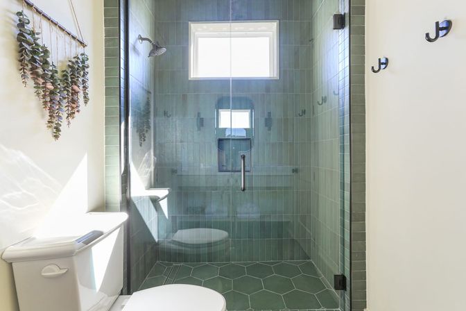 Sunlit modern bathroom with glass-enclosed walk-in shower clad in vertical green tiles and matching hexagon floor tiles, matte black hardware, white toilet, and hanging eucalyptus wall decor.