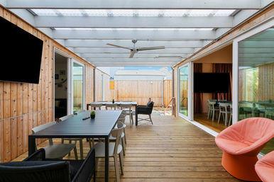 Sunlit residential covered wooden patio with long outdoor dining table, mixed modern chairs, ceiling fan, wall-mounted flat-screen TV, sliding glass doors to interior rooms, and cozy coral accent chairs overlooking a fenced backyard.