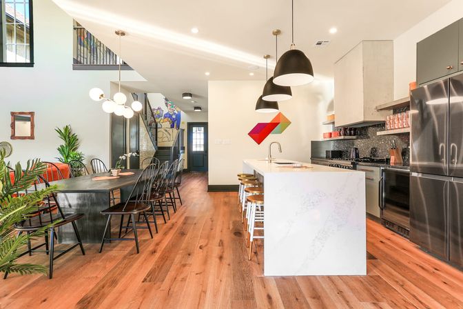 Bright modern open-concept kitchen and dining room with white marble island, black pendant lights, stainless appliances, long wooden dining table, hardwood floors, indoor plants and colorful geometric wall art.