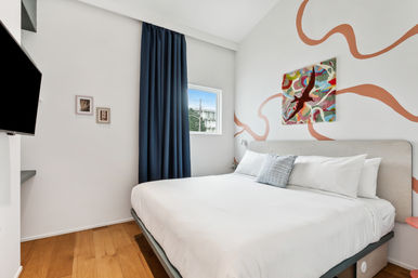Bright modern minimalist bedroom with white king bed, colorful abstract wall art and coral mural accents, navy curtains, small window and hardwood floor