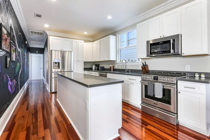 Bright modern white kitchen with gray island countertop, stainless-steel refrigerator, range and microwave, glossy hardwood floors and a chalkboard-art mural along the hallway wall