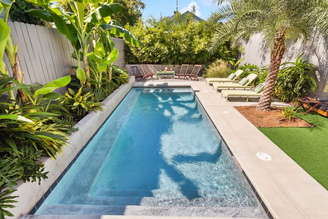 Sun-drenched narrow rectangular backyard pool with clear blue water, concrete deck, tropical palms and banana plants, green lounge chairs and wooden folding chairs at the far end.