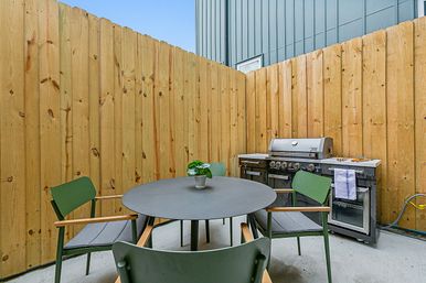 Cozy fenced patio with a round black dining table, four green chairs, small potted plant centerpiece and a stainless steel gas grill against a tall wooden privacy fence