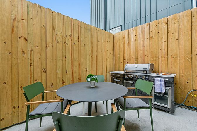 Cozy fenced patio with a round black dining table, four green chairs, small potted plant centerpiece and a stainless steel gas grill against a tall wooden privacy fence