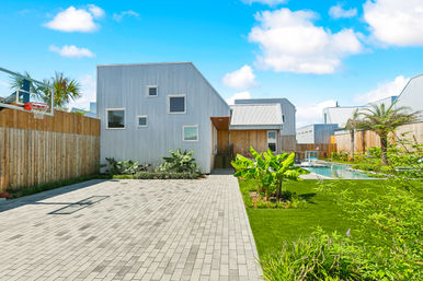 Sunny modern coastal house with corrugated metal siding, paved driveway and basketball hoop, lush tropical landscaping and green lawn leading to a backyard swimming pool under a bright blue sky.