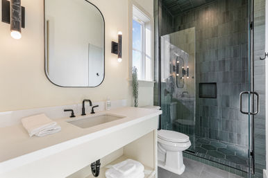 Bright, modern bathroom with white vanity and matte black fixtures, glass-enclosed gray-tile walk-in shower with hex floor tiles, wall mirror and window letting in natural light.