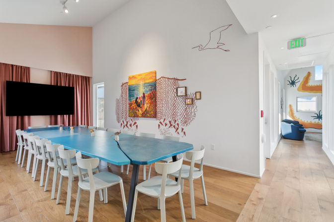 Bright coastal-themed meeting room with a long blue modular conference table, white chairs, wood floors, pink curtains, wall-mounted TV, seascape painting framed by net-style wall art, and a hallway featuring a colorful mural and blue lounge chairs.