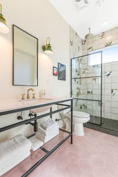 Bright beach-inspired bathroom with pink hexagon tile floor, black-framed open vanity with folded white towels, brass faucet, green wall sconces, and glass walk-in shower with tropical bird-and-leaf tiles