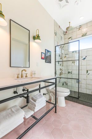 Bright beach-inspired bathroom with pink hexagon tile floor, black-framed open vanity with folded white towels, brass faucet, green wall sconces, and glass walk-in shower with tropical bird-and-leaf tiles