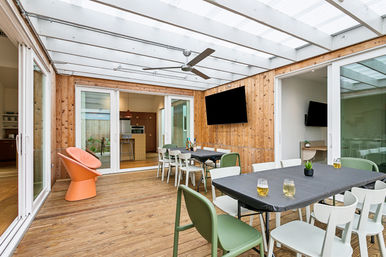 Bright enclosed sunroom dining area with glass roof and wood-paneled walls, ceiling fan, wall-mounted TV, long tables with mixed chairs and drinks, and an orange lounge chair.