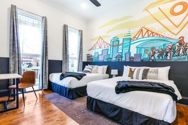 Bright urban boutique hotel room with two double beds, hardwood floors, large windows with patterned curtains, desk and leather chair, and a colorful city skyline and bridge mural above the headboards.