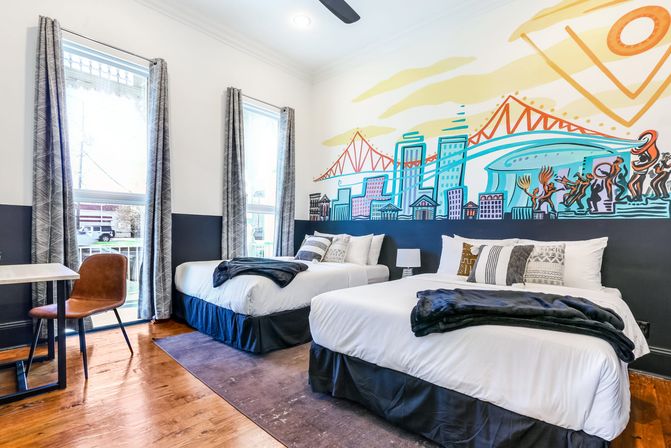 Bright urban boutique hotel room with two double beds, hardwood floors, large windows with patterned curtains, desk and leather chair, and a colorful city skyline and bridge mural above the headboards.