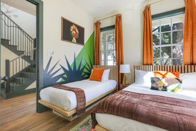Sunlit vacation-rental bedroom with two platform beds (single and double), striped headboards, orange curtains framing windows with tree views, green geometric wall mural, colorful throw pillows and brown blankets on hardwood floors, staircase visible through open doorway.